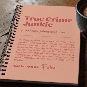 May include: A spiral-bound notebook with the text "True Crime Junkie" in a salmon-colored font. The notebook is open, revealing a definition of the term. A cup of coffee with latte art sits next to the notebook.