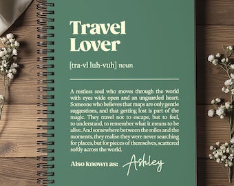 May include: A green notebook with a spiral binding, displaying the words "Travel Lover" in white. The definition of "Travel Lover" is printed below the title. The notebook rests on a wooden surface, with white flowers and fabric.