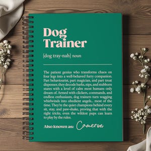 Może przedstawiać: Spiralny notatnik z turkusową okładką z różowym napisem "Dog Trainer". Poniżej tytułu wydrukowano definicję trenera psów. Notatnik leży na drewnianej powierzchni z kwiatowymi akcentami.