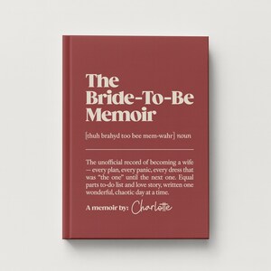 May include: A dark red hardcover book titled The Bride-To-Be Memoir in cream-colored serif font. The cover includes a phonetic pronunciation guide, the noun classification, and a descriptive paragraph about wedding planning, signed by Charlotte.