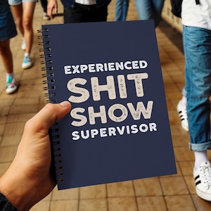 Peut inclure: Un cahier bleu marine à spirales avec l'inscription "EXPERIENCED SHIT SHOW SUPERVISOR" en lettres blanches texturées. Le cahier est tenu dans une main, avec un arrière-plan flou de personnes et un sol carrelé.