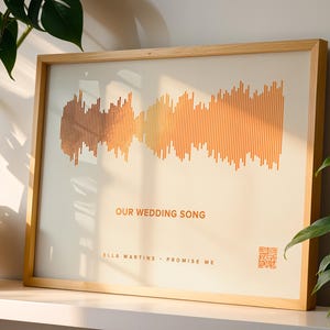 May include: A framed print featuring a soundwave design in shades of orange and brown. The text "OUR WEDDING SONG" is displayed, along with artist and song title. The frame is made of light-colored wood, and a QR code is visible.