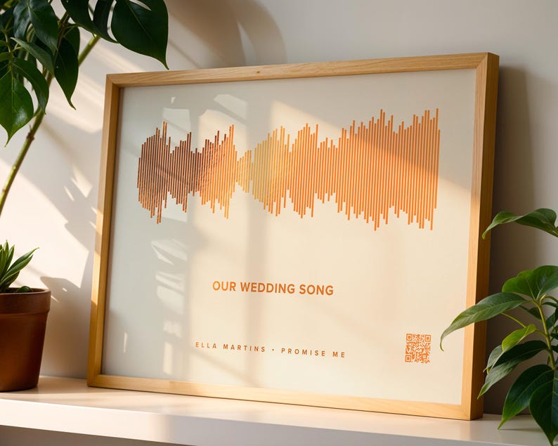 May include: A framed print featuring a soundwave design in shades of orange and brown. The text "OUR WEDDING SONG" is displayed, along with artist and song title. The frame is made of light-colored wood, and a QR code is visible.