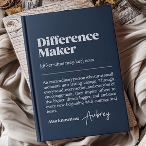 Puede incluir: Un diario de tapa dura azul marino con las palabras "Difference Maker" en blanco. El diario incluye una definición y el nombre "Aubrey" en escritura cursiva. El diario está sobre una superficie de madera.