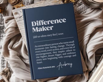 May include: A navy blue hardcover journal with the words "Difference Maker" in white. The journal includes a definition and the name "Aubrey" in script. The journal is resting on a wooden surface.