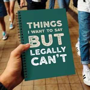 Könnte beinhalten: Ein türkisgrünes Spiralnotizbuch mit dem Text "THINGS I WANT TO SAY BUT LEGALLY CAN'T" in Weiß. Das Notizbuch wird von einer Person in der Hand gehalten, mit anderen Personen im Hintergrund.