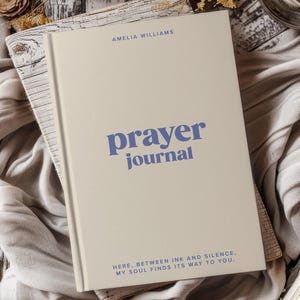 May include: A beige hardback prayer journal with blue text. The cover features the name Amelia Williams at the top, the title prayer journal in the centre, and the phrase Here, between ink and silence, my soul finds its way to you.