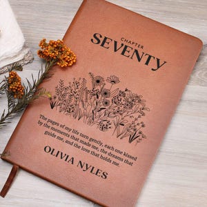 May include: A brown leather journal with the text "CHAPTER SEVENTY" and a floral design. The quote "The pages of my life turn gently, each one kissed by the moments that made me, the dreams that guide me, and the love that holds me." is printed below the floral design. The name "OLIVIA NYLES" is printed at the bottom of the journal.