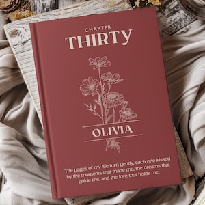 Puede incluir: Un libro granate con el texto "CHAPTER THIRTY" y el nombre "OLIVIA" en blanco. Una ilustración floral está centrada en la portada. El libro tiene una cita en la parte inferior. El libro está sobre una superficie de madera.
