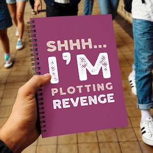 May include: A magenta spiral-bound notebook with the text "SHHH... I'M PLOTTING REVENGE" in decorative fonts. The notebook is held in a person's hand. The background shows people walking on a paved surface.