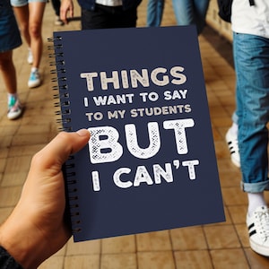 以下が含まれることがあります： 「THINGS I WANT TO SAY TO MY STUDENTS BUT I CAN'T」の文字が白とベージュで書かれた、ネイビーブルーのリング綴じノート。ノートは人の手で持たれており、背景には他の人がいます。