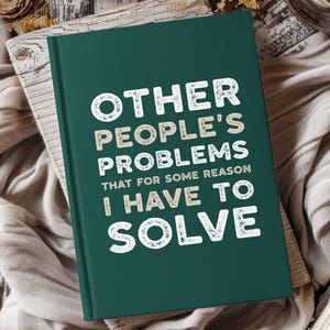 以下が含まれることがあります： 「OTHER PEOPLE'S PROBLEMS THAT FOR SOME REASON I HAVE TO SOLVE」の文字が白とベージュで書かれた、濃いティール色のジャーナル。ジャーナルは、ニュートラルカラーの布が敷かれた木製の表面に置かれています。