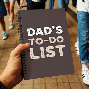 Może przedstawiać: Ciemnoszary notatnik w spiralę z białym, teksturowanym napisem "DAD'S TO-DO LIST". Notatnik trzymany jest w dłoni osoby, w tle widać ludzi i drewnianą podłogę.