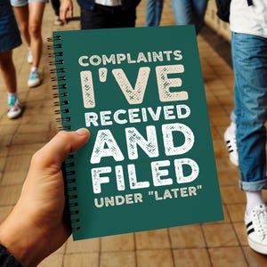 May include: A dark teal spiral-bound notebook featuring the white text COMPLAINTS I'VE RECEIVED AND FILED UNDER "LATER" printed on the front cover. The notebook has a black metal wire binding along the left side and a matte finish on the cover.