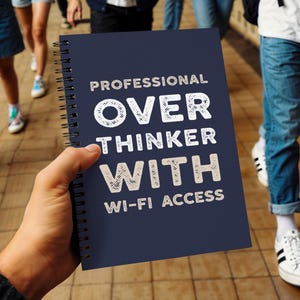 Peut inclure: Un cahier à spirales bleu marine avec le texte "PROFESSIONNEL OVER THINKER WITH WI-FI ACCESS" en blanc. Le cahier est tenu dans la main d'une personne, avec des personnes et un sol carrelé en arrière-plan.