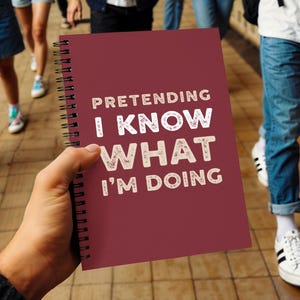 以下が含まれることがあります： 「PRETENDING I KNOW WHAT I'M DOING」の白い文字が書かれた、ワインレッドのリングノート。人が持っています。背景には、舗装された表面を歩く人々がいます。