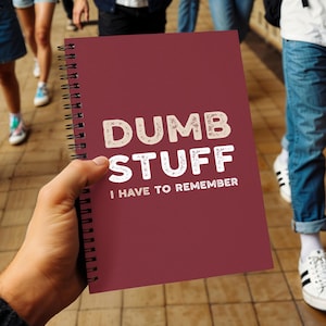 Può includere: Un taccuino a spirale bordeaux con la scritta "DUMB STUFF I HAVE TO REMEMBER" in bianco e bianco sporco. Il taccuino è tenuto in mano, con persone e un pavimento piastrellato sullo sfondo.