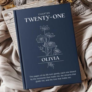 May include: A dark blue hardcover book featuring the text "CHAPTER TWENTY-ONE" at the top, a white line-art illustration of a floral bouquet in the center, the name "OLIVIA" below the flowers, and a poetic quote printed at the bottom.