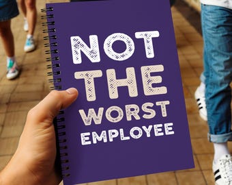 May include: A purple spiral-bound notebook with the text "NOT THE WORST EMPLOYEE" in white. The notebook is held in a person's hand, with other people and legs visible in the background.