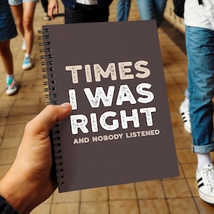 May include: A dark gray spiral-bound notebook with the text "TIMES I WAS RIGHT AND NOBODY LISTENED" in white and beige lettering. The notebook is held in front of a blurred background of people walking.