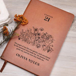 Puede incluir: Un diario de cuero marrón con el texto "CHAPTER 21" y un diseño floral. La cita "The pages of my life turn gently, each one kissed by the moments that made me, the dreams that guide me, and the love that holds me." está impresa en la portada. El nombre "OLIVIA NYLES" también está impreso en la portada.