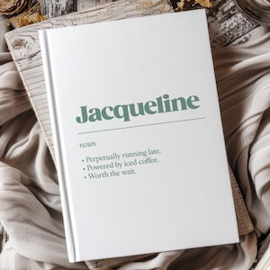 May include: A white hardback book featuring the name Jacqueline in large green text, followed by the noun definition: Perpetually running late. Powered by iced coffee. Worth the wait. The text is clearly printed on the front cover of the book.