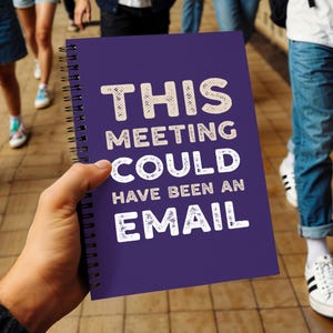 May include: A purple spiral-bound notebook with the text "THIS MEETING COULD HAVE BEEN AN EMAIL" in white, textured lettering. The notebook is held by a person, with a blurred background of people and a tiled floor.