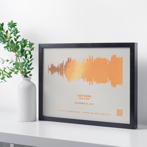 May include: Framed artwork featuring a soundwave design in shades of orange and gold. The text "OUR SONG" is displayed, along with names and a date. The artwork is in a black frame and is a decorative piece.