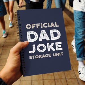 Op de afbeelding: Een marineblauw spiraalgebonden notitieboek met de tekst "OFFICIAL DAD JOKE STORAGE UNIT" in witte en beige letters. Het notitieboek wordt in de hand van een persoon gehouden, met mensen die op de achtergrond lopen.