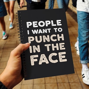 Op de afbeelding: Zwart spiraalgebonden notitieboek met de tekst "PEOPLE I WANT TO PUNCH IN THE FACE" in witte en beige letters. Het notitieboek wordt in de hand van een persoon gehouden, met mensen en een betegelde vloer op de achtergrond.