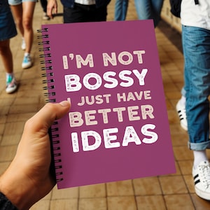 Op de afbeelding: Een spiraalgebonden notitieboek met een diep magenta omslag. Het notitieboek bevat de tekst "I'M NOT BOSSY I JUST HAVE BETTER IDEAS" in een witte, getextureerde letter. Het notitieboek wordt in een hand gehouden.