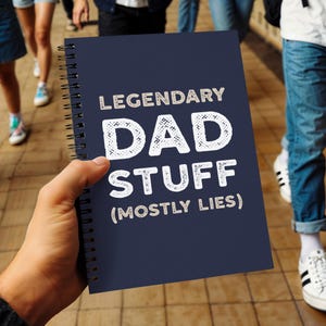 May include: A navy blue spiral-bound notebook with the text "LEGENDARY DAD STUFF (MOSTLY LIES)" in white lettering. The notebook is held in a person's hand, with other people and a tiled floor in the background.