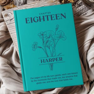 May include: A teal-colored book with the text "CHAPTER EIGHTEEN" and "HARPER" in dark teal. A floral illustration is below the text. The book has a quote at the bottom. The book is on a wooden surface with a gray cloth.