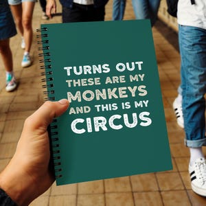 May include: A teal spiral-bound notebook with the text "TURNS OUT THESE ARE MY MONKEYS AND THIS IS MY CIRCUS" in white. The notebook is held in front of a blurred background of people walking.
