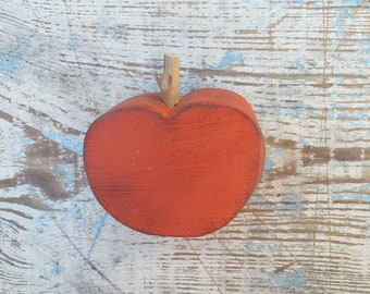 May include: A red wooden apple with a twig stem. The apple is sitting on a whitewashed wooden surface.
