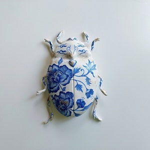 Könnte beinhalten: Ein weißer Keramikkäfer mit blauen Blumenmustern. Der Käfer hat sechs Beine und ein detailliertes Blumenmuster auf dem Rücken.
