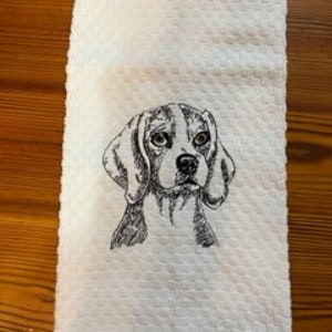 May include: White, textured hand towel featuring an embroidered black and white Beagle dog portrait. The towel has a waffle weave pattern and is folded neatly. The Beagle's face is centered, showcasing its floppy ears and soulful eyes.