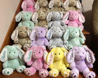 May include: A collection of plush bunny rabbits in various pastel colors, including pink, blue, and yellow. Each bunny has long ears with embroidered names and dates. The bunnies are arranged on wooden steps.