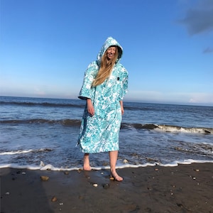 Puede incluir: Una persona lleva una toalla con capucha turquesa y blanca con un estampado floral. La toalla se lleva sobre un traje de baño y la persona está de pie en una playa de arena.