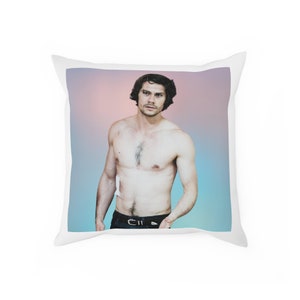 May include: A shirtless man with dark hair and a muscular build stands against a pink and blue gradient background. He is wearing black jeans and a black belt.