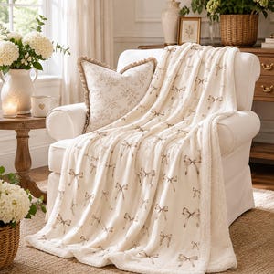 May include: Cream-colored throw blanket with a repeating pattern of brown bows, draped over a white armchair. The blanket has a soft, plush texture and a sherpa lining. A decorative pillow with a floral design rests on the chair.