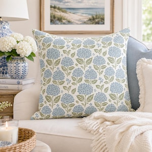 May include: A square throw pillow with a floral pattern of blue hydrangeas and green leaves on a cream background. The pillow is placed on a white sofa, alongside other decorative pillows and a white blanket.