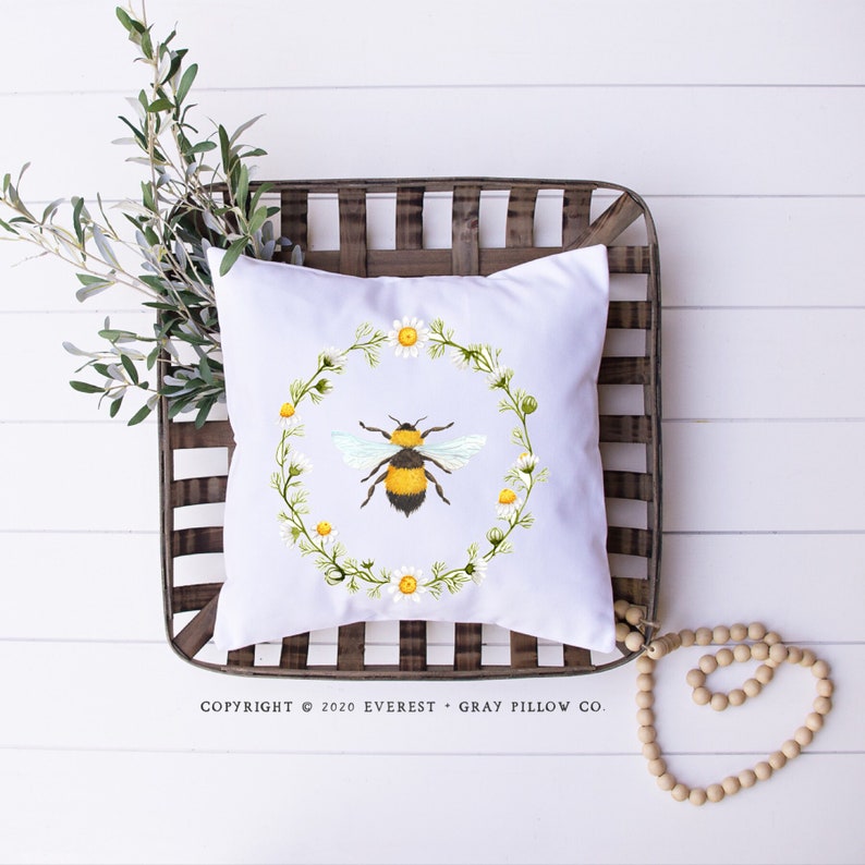 Puede incluir: Coj&iacute;n cuadrado blanco con una ilustraci&oacute;n de acuarela de una abeja dentro de una corona de margaritas y vegetaci&oacute;n. El coj&iacute;n est&aacute; colocado en una cesta de madera r&uacute;stica sobre una superficie de madera blanca. Texto: "COPYRIGHT &copy; 2020 EVEREST &bull; GRAY PILLOW CO."