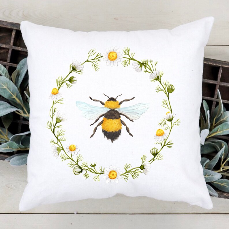 Puede incluir: Coj&iacute;n cuadrado blanco con una ilustraci&oacute;n de acuarela de un abejorro amarillo y negro, rodeado por una corona de margaritas y hojas verdes. Un art&iacute;culo decorativo para el hogar.