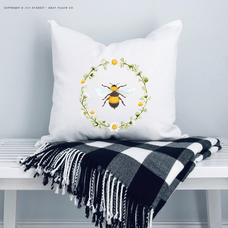Puede incluir: Coj&iacute;n cuadrado blanco con un dise&ntilde;o de acuarela de una abeja dentro de una corona de margaritas. El coj&iacute;n est&aacute; sobre un banco blanco, acompa&ntilde;ado de una manta a cuadros en blanco y negro con flecos. Copyright 2020 Everest Gray Pillow Co.