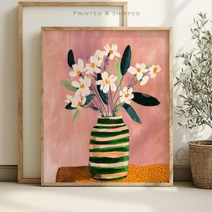 May include: A framed art print featuring a painting of white flowers with yellow centers in a green and peach striped vase. The vase sits on a surface with a yellow and black polka dot pattern against a soft, textured pink background.