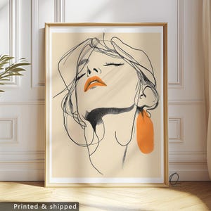 Peut inclure: Une impression d'art encadrée représentant un dessin au trait minimaliste d'un visage de femme aux yeux fermés et aux lèvres orange. L'œuvre comprend une boucle d'oreille orange et est sur fond beige. Le cadre est doré.