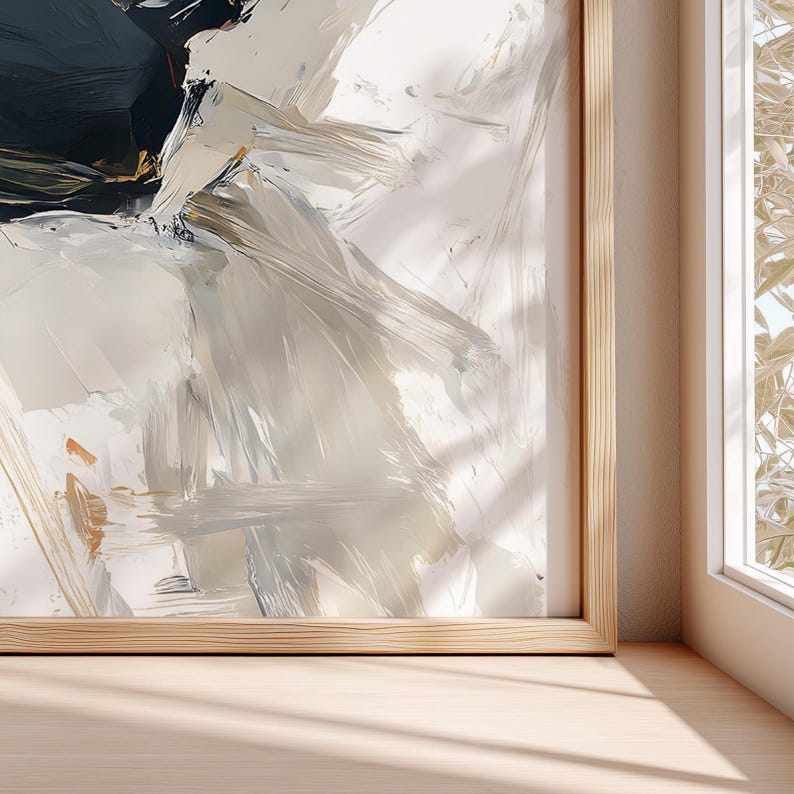 May include: Abstract art print with a wooden frame. The artwork features broad brushstrokes in shades of white, beige, and navy blue. The painting is displayed on a light-colored wooden surface, with a window and sunlight visible in the background.