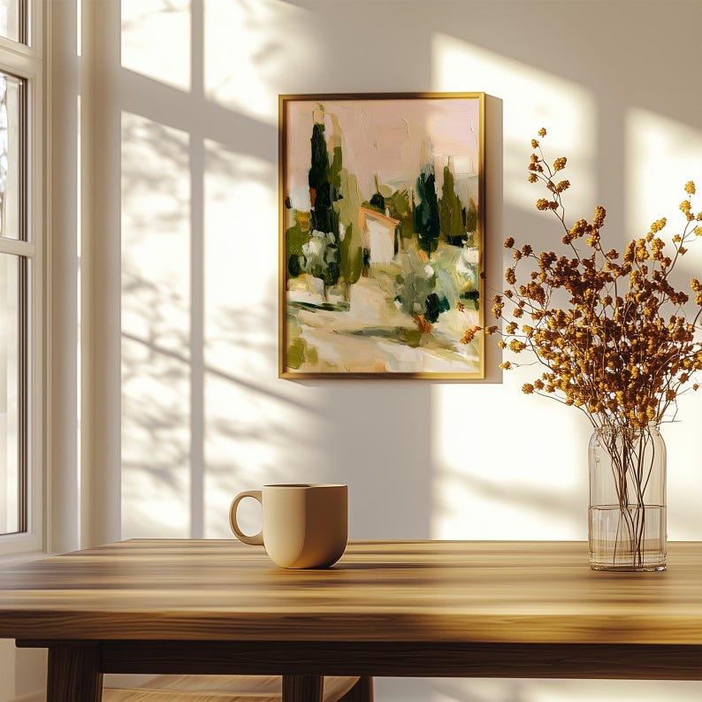 Puede incluir: Un cuadro de paisaje enmarcado con cipreses verdes abstractos y un peque&ntilde;o edificio sobre un cielo rosa suave. Una taza de cer&aacute;mica beige lisa y un jarr&oacute;n de cristal transparente con flores secas marrones sobre una mesa de madera.