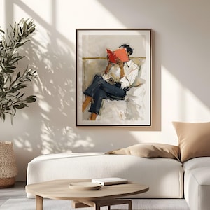 May include: Framed art print featuring an abstract painting of a person reading a book. The person is wearing a white shirt and dark blue pants. The book is bright red, and the artwork has a neutral color palette with a gold frame.